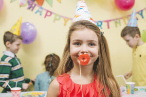 Blog 12 Girl at a birthday party wearing a party hat and playful red lips decoration, surrounded by festive decorations and children celebrating.
