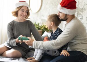 Blog 8 Family celebrating Christmas together, wearing festive Santa hats, with a woman handing a gift to a young boy while a man watches, capturing the warmth of holiday gatherings and family moments.