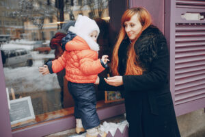Blog 7 Mother helping child in winter clothing reach a shop window, showcasing family engagement and joyful moments during the holiday season.