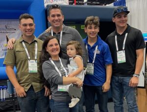 Blog 6 Group of six individuals, including a woman holding a child, posing together at a convention or event, showcasing community engagement in the theme park industry.